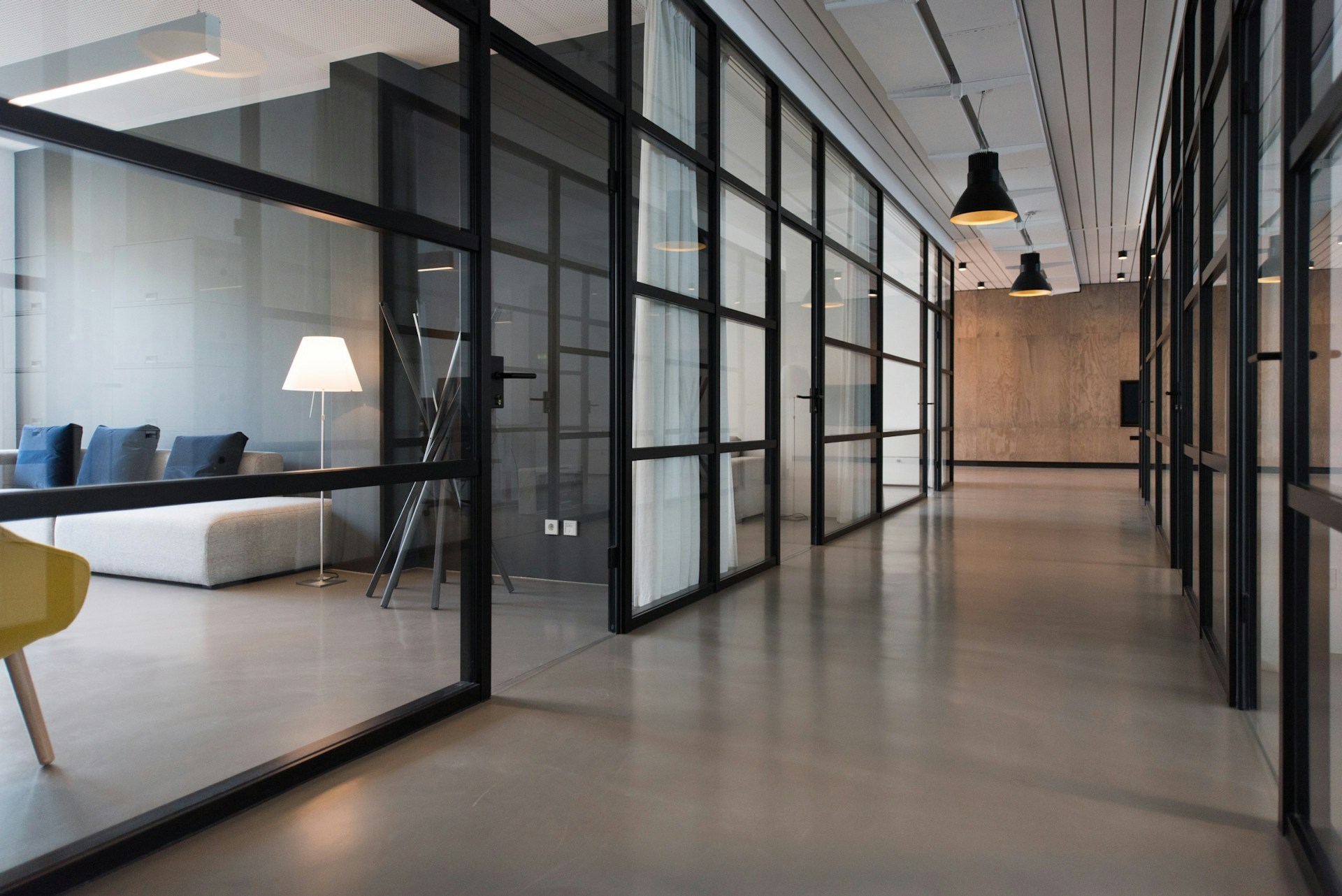 Glasreinigung Das Bild zeigt einen modernen Bürogang mit großen Glaswänden, die von schwarzen Rahmen gehalten werden. Links sind durch die Glaswand Möbel in einem Büro zu sehen: ein cremefarbenes Sofa mit blauen Kissen und eine weiße Stehlampe mit einem hellen Schirm. Ein gelber Stuhl ist im Vordergrund links nur teilweise sichtbar. Der Boden des Ganges ist glänzend grau und reflektiert die Deckenbeleuchtung. An der Decke des Ganges hängen schwarze, glockenförmige Lampen. Der Gang erstreckt sich in die Ferne, und die Glaswände setzen sich fort, was den Eindruck eines großen, offenen und gleichzeitig unterteilten Raumes vermittelt. Das gesamte Bild strahlt eine schlichte, moderne Ästhetik aus.