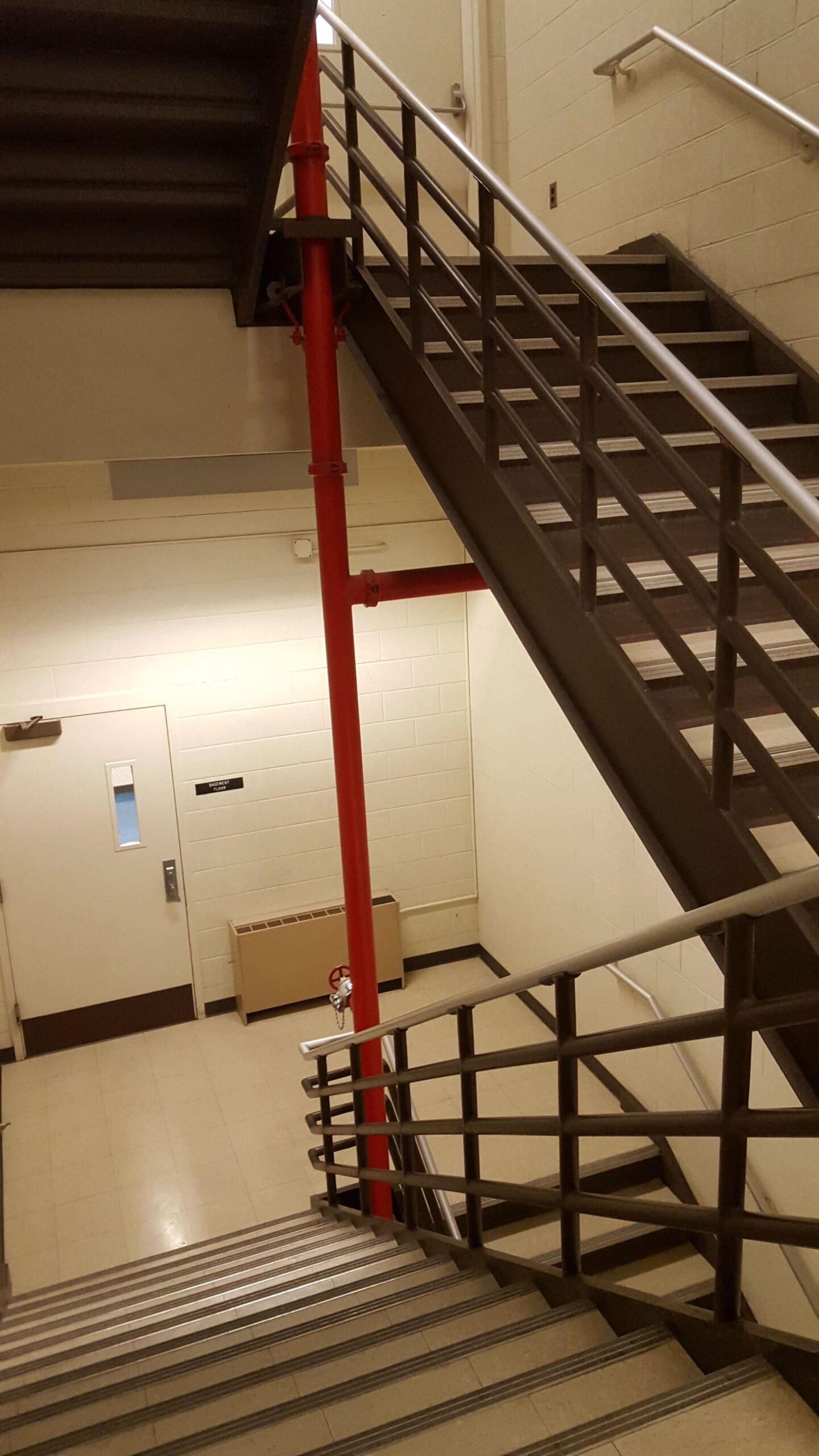 Treppenhausreinigung Das Bild zeigt einen Innenbereich mit einem Treppenhaus, das aus zwei sich kreuzenden Treppenläufen besteht. Die Treppen sind aus Metall gefertigt, mit dunkelbraunen Stufen und einem Handlauf aus hellem Metall. In der Mitte des Bildes und zwischen den Treppenläufen verläuft ein auffälliges rotes Rohr, das Teil eines Sprinklersystems oder einer Feuerlöschleitung zu sein scheint. Von diesem vertikalen Rohr zweigen horizontale rote Rohre ab. Im Hintergrund ist eine helle Wand mit einer weißen Tür und einem kleinen Fenster zu sehen. Unterhalb des horizontalen roten Rohrs an der Wand befindet sich ein beiger Heizkörper oder eine Lüftungsanlage. Der Boden im Treppenhaus ist hell und reflektiert das Licht. Die Beleuchtung ist hell, was den Raum gut ausleuchtet. Das Gesamtbild vermittelt den Eindruck eines funktionalen und sicherheitsorientierten Gebäudebereichs, möglicherweise in einem öffentlichen Gebäude oder einer Industrieanlage.