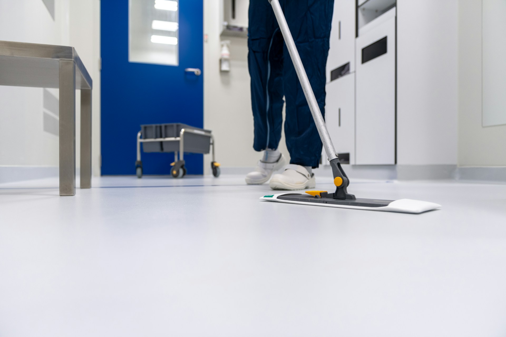 Büroreinigung Das Bild zeigt einen Mann in blauer Hose und weißen Schuhen, der in einem hellen, sauberen Raum einen weißen Wischmopp über einen glänzenden, hellgrauen Boden schiebt. Ein Teil des silbernen Griffs des Wischmopps ist sichtbar, und der orangefarbene Griffkopf befindet sich in der Mitte des Bildes. Im Hintergrund ist ein Teil eines Edelstahl- oder Metalltisches zu sehen. Auf der rechten Seite sind weiße Schränke erkennbar. Eine blaue Tür mit einem Fenster ist ebenfalls im Hintergrund auf der linken Seite zu sehen. Ein grauer Rollwagen mit einem Eimer oder Behälter steht in der Nähe der blauen Tür. Das Licht im Raum ist hell, und die gesamte Umgebung wirkt sehr steril und sauber.